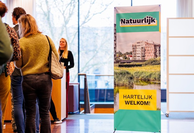 Mensen staan in een rij bij een ontvangstbalie naast een banner met de tekst “Natuurlijk” en “Hartelijk welkom!”