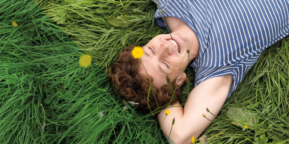 Persoon ligt glimlachend in het gras met bloemen om zich heen.