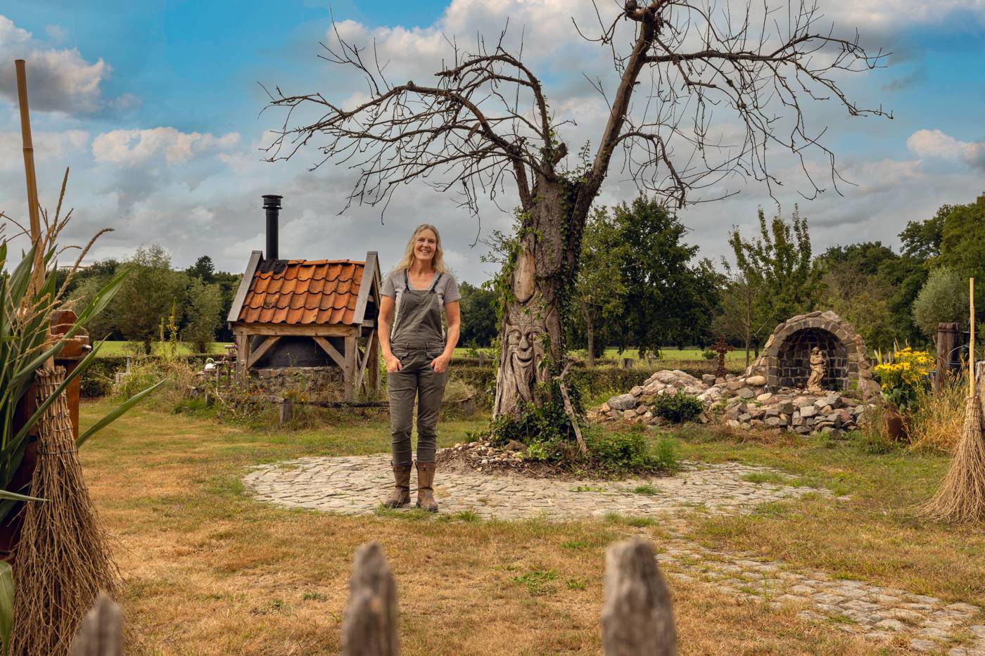 Vrouw staat in een tuin bij een boom met houtsnijwerk en een klein gebouw met rood dak.