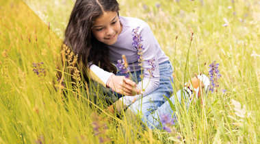 Meisje knielt in een bloemenweide en bekijkt paarse bloemen tussen hoog gras.