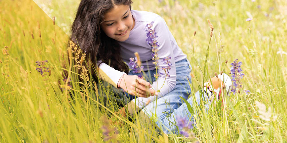 Meisje knielt in een bloemenweide en bekijkt paarse bloemen tussen hoog gras.