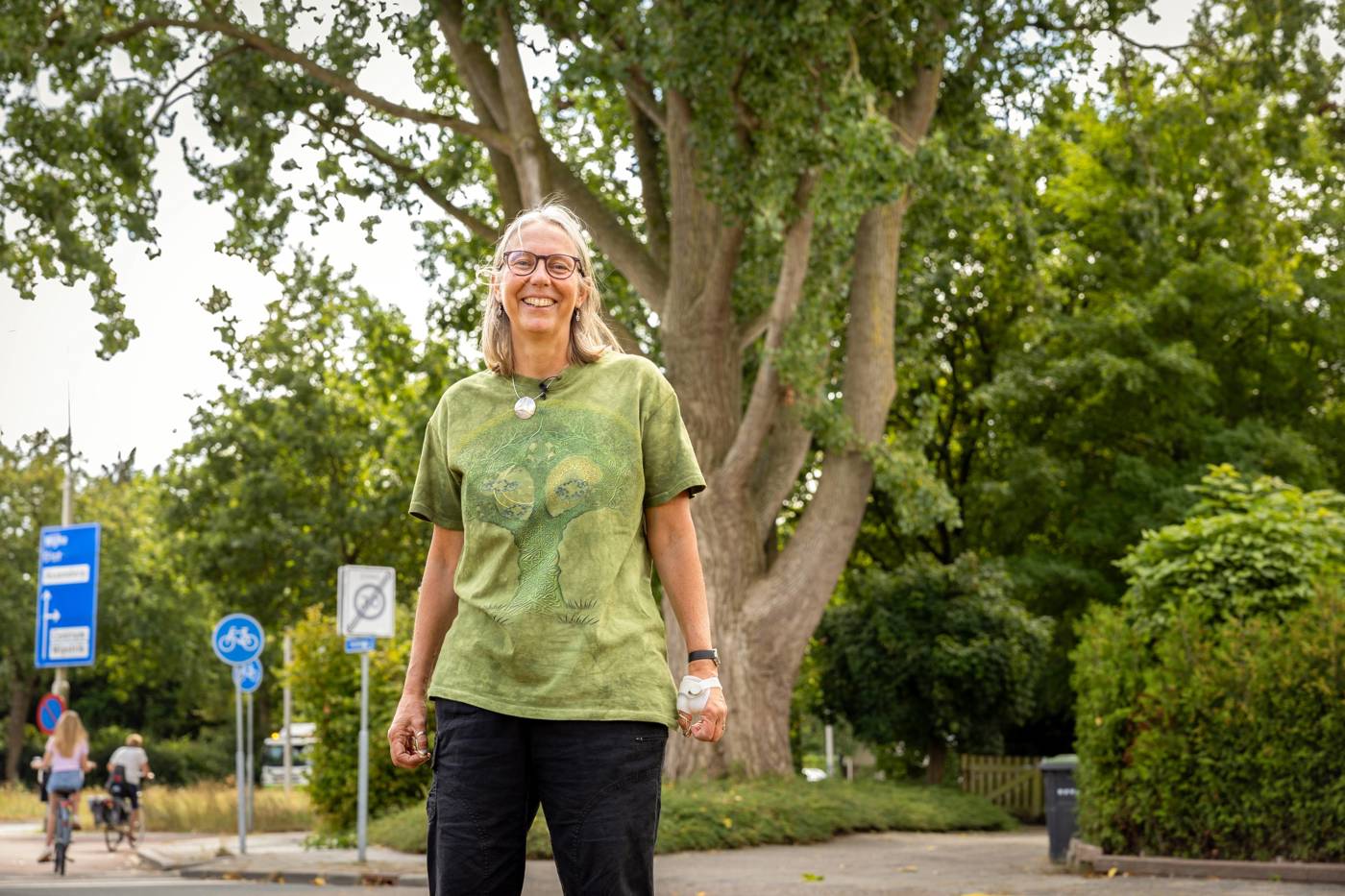 Vrouw staat lachend op straat met bomen en verkeersborden op de achtergrond.