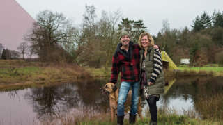 Man en vrouw staan met hun hond bij een vijver in een groen landschap met bomen en een tent op de achtergrond.