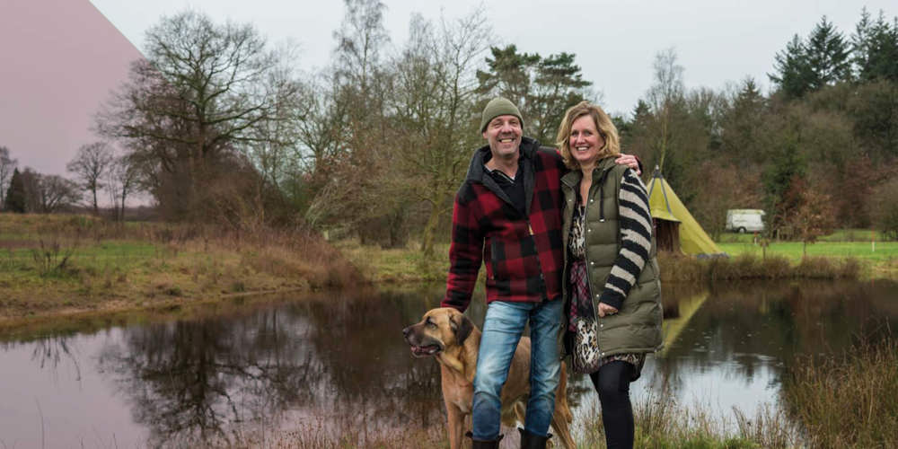 Man en vrouw staan met hun hond bij een vijver in een groen landschap met bomen en een tent op de achtergrond.