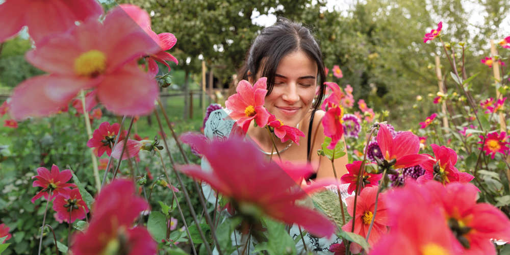 Vrouw glimlacht tussen felroze bloemen in een tuin op een zomerse dag.