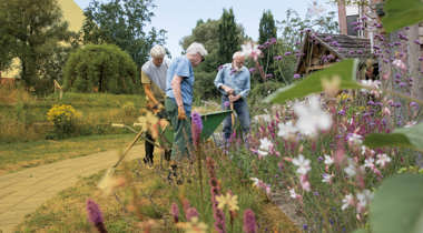Drie personen werken in een tuin met bloemen en planten, gebruikmakend van tuingereedschap en een kruiwagen.
