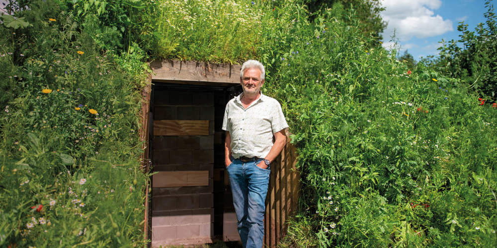 Een man staat voor een houten deur in een groene heuvel, omringd door wilde planten