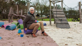 Een vrouw zit op een speeltoestel in een zandbak met kleurrijk speelgoed en bomen op de achtergrond.