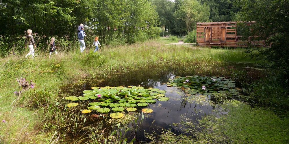 Een gezin loopt langs een vijver met waterlelies in een groene, natuurlijke omgeving met een houten gebouw op de achtergrond.