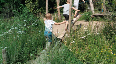 Drie kinderen spelen op houten structuren in een groene omgeving.
