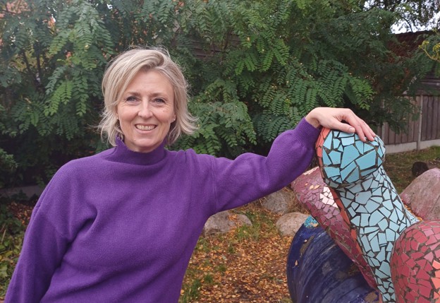 Vrouw in paarse trui leunt lachend tegen een kleurrijk mozaïekbeeld in een tuin.