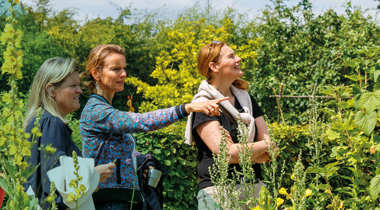 Drie personen staan in een groene tuin met bloeiende planten, terwijl één persoon naar iets wijst.